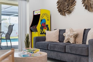 A living room with a grey sofa, a yellow arcade machine, a starfish pillow, and a small table with a plant and books.
