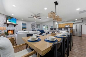A modern kitchen with stainless steel appliances, a dining area with a wooden table set for six, and a living room with a fireplace and a large painting of a beach scene.