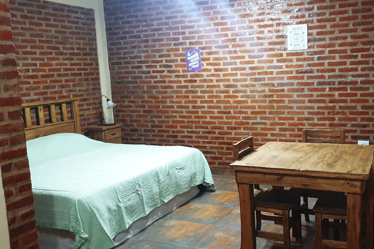 A room with a brick wall, a bed with a green bedspread, a wooden table with chairs, and a nightstand with a lamp.