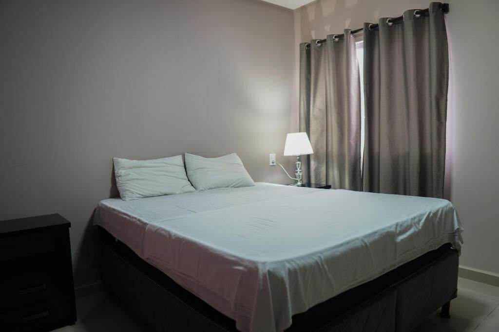A single bed with a white mattress and pillows, a bedside table with a lamp, and a window with curtains.