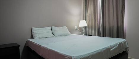 A single bed with a white mattress and pillows, a bedside table with a lamp, and a window with curtains.