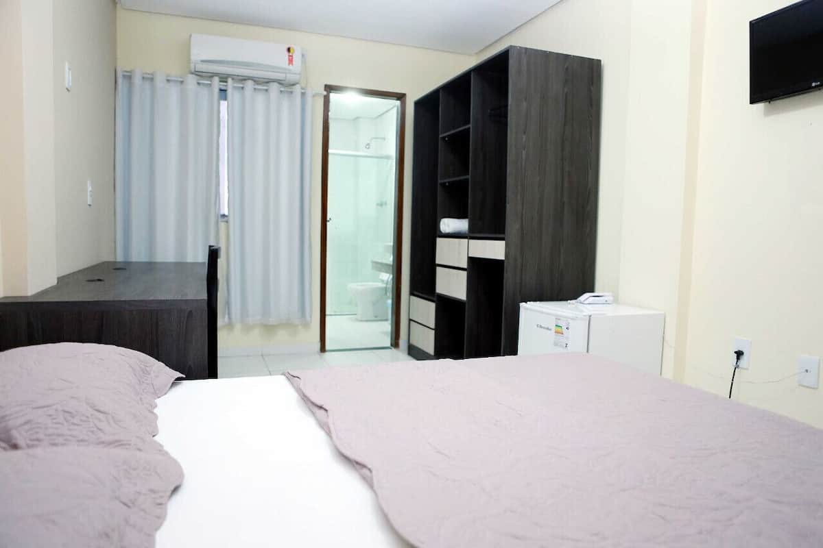 A bedroom with a bed, a desk, a wardrobe, a small white appliance, and a bathroom visible through a glass door.