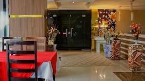 Un hall de hotel con mostrador de recepción, una mesa con cubierta roja y una silla. Hay jarrones decorativos con flores y suelo de baldosas.