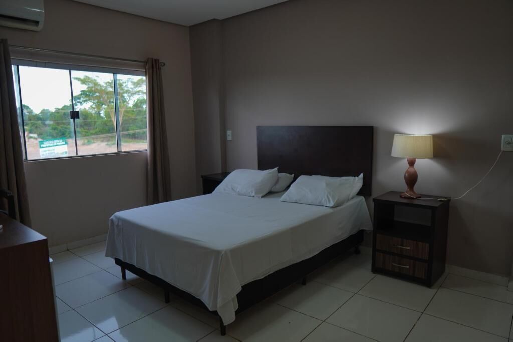 A bedroom with a bed, a nightstand, a lamp, a window with curtains, and a view of trees outside.