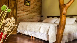 A bedroom with a brick wall, a wooden door, a bed with white bedding, and a tree trunk in the foreground. - La Maina Cabañas (San Javier)