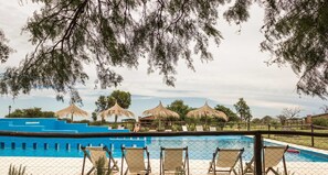 A pool area with lounge chairs and thatched umbrellas, surrounded by a fence and trees.