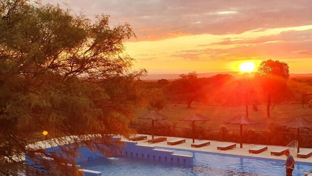 A swimming pool with lounge chairs and umbrellas, surrounded by trees and a sunset in the background. - La Maina Cabañas (San Javier)