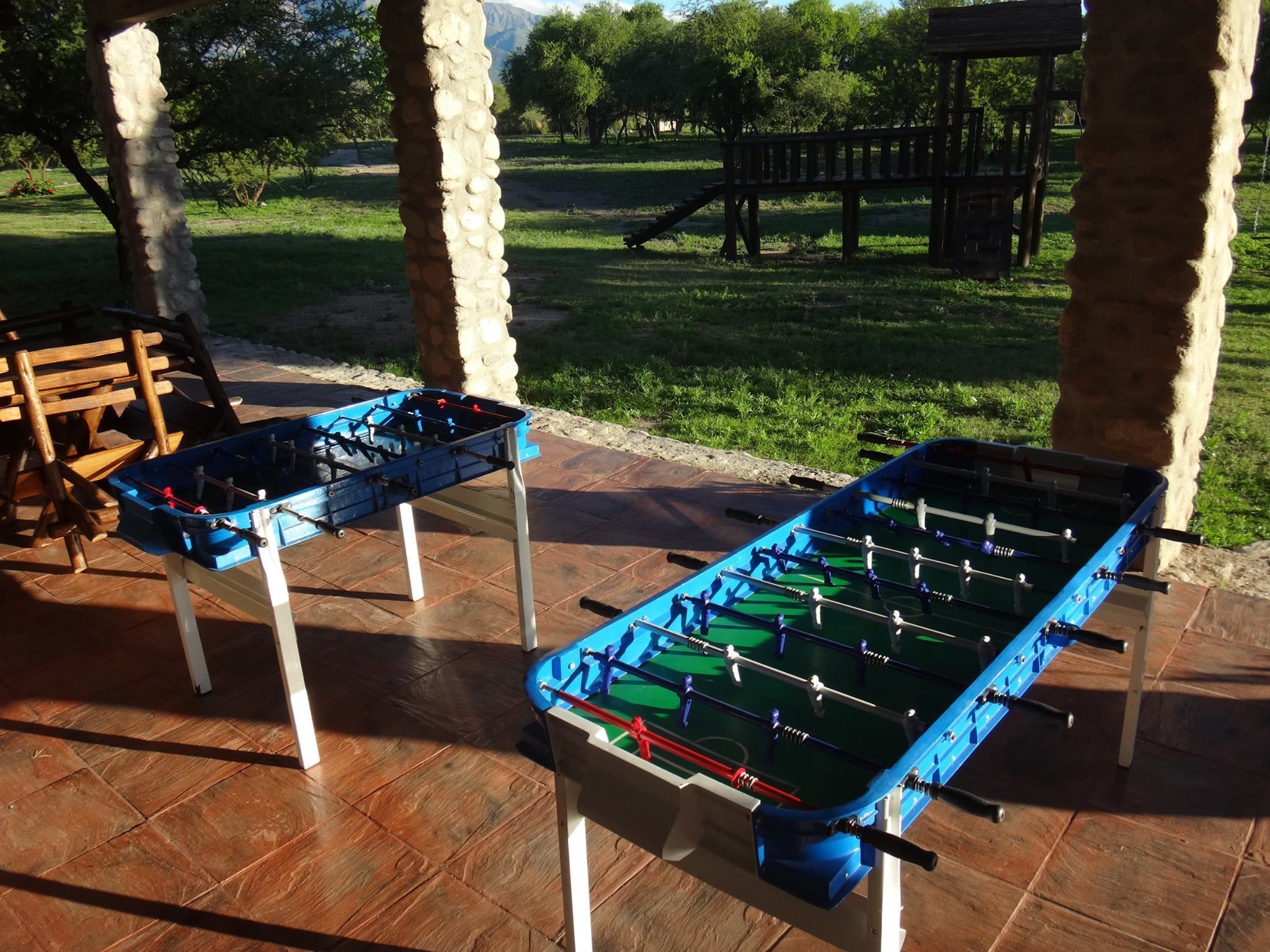 Dos mesas de futbolín instaladas al aire libre en un área techada, con un campo de césped y árboles al fondo.