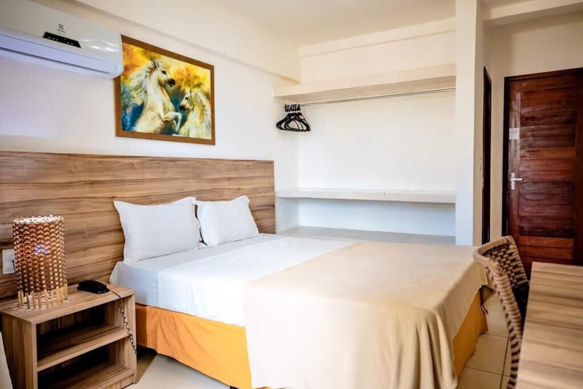A hotel room with a wooden headboard, a bed with white bedding, a painting of horses on the wall, a coat rack, and a wooden door.