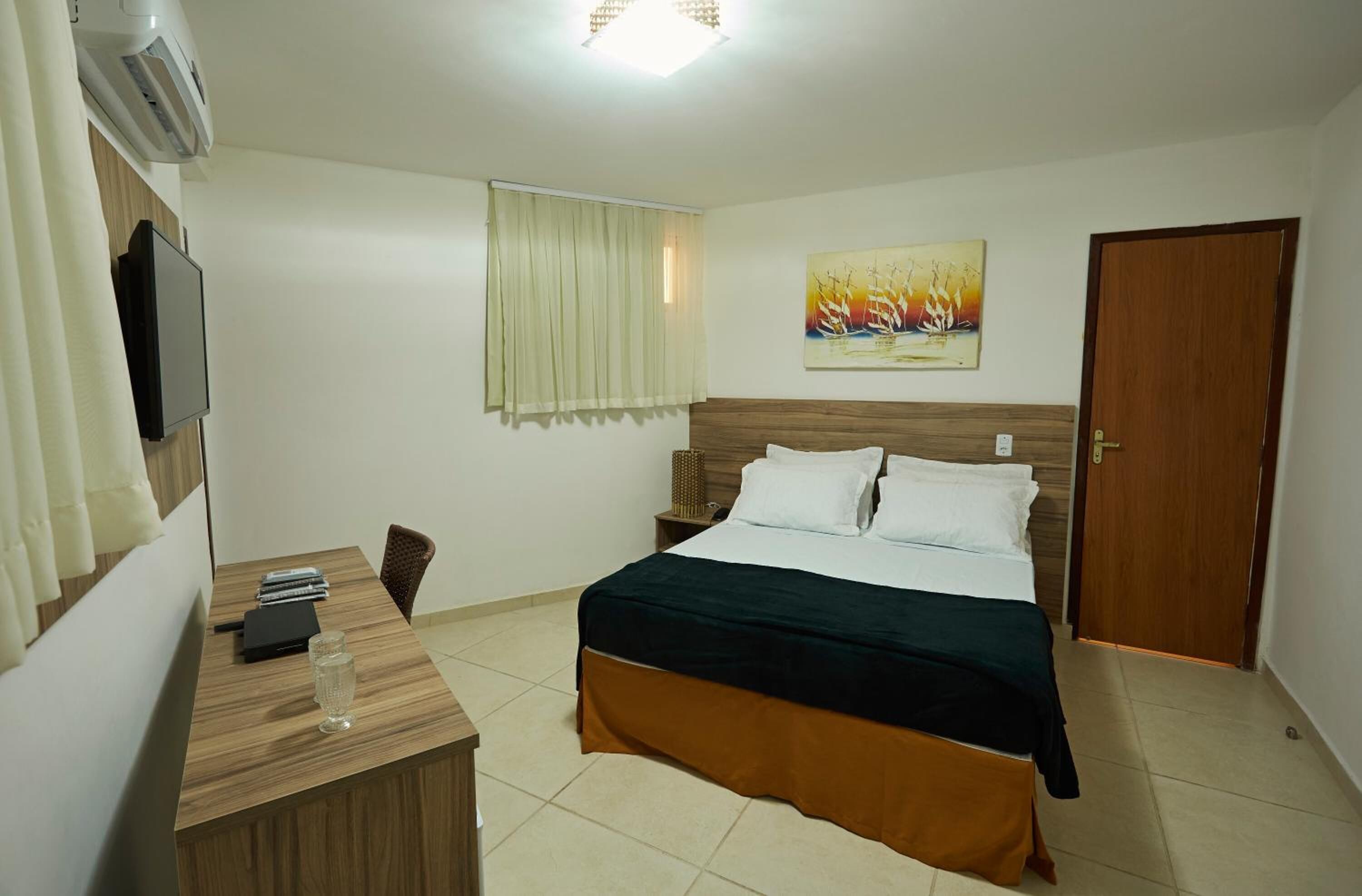 Una habitación de hotel con una cama, un escritorio con computadora, un televisor y un cuadro en la pared.