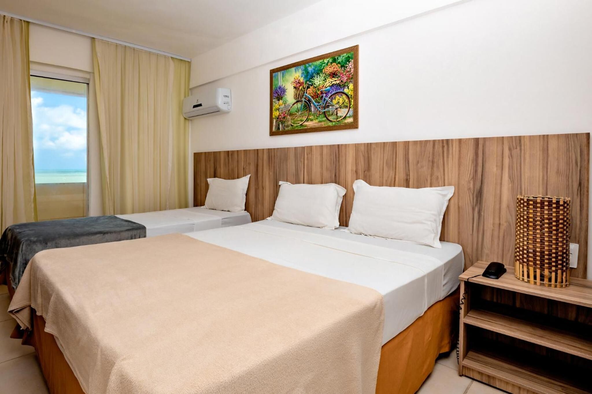 Une chambre d’hôtel avec un grand lit, deux tables de chevet, une tête de lit en bois, un tableau représentant un vélo et une vue sur la plage par la fenêtre.