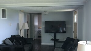 Una sala de estar con dos sofás de cuero negro, una mesa de centro de madera, un televisor montado en la pared y un ventilador de techo.