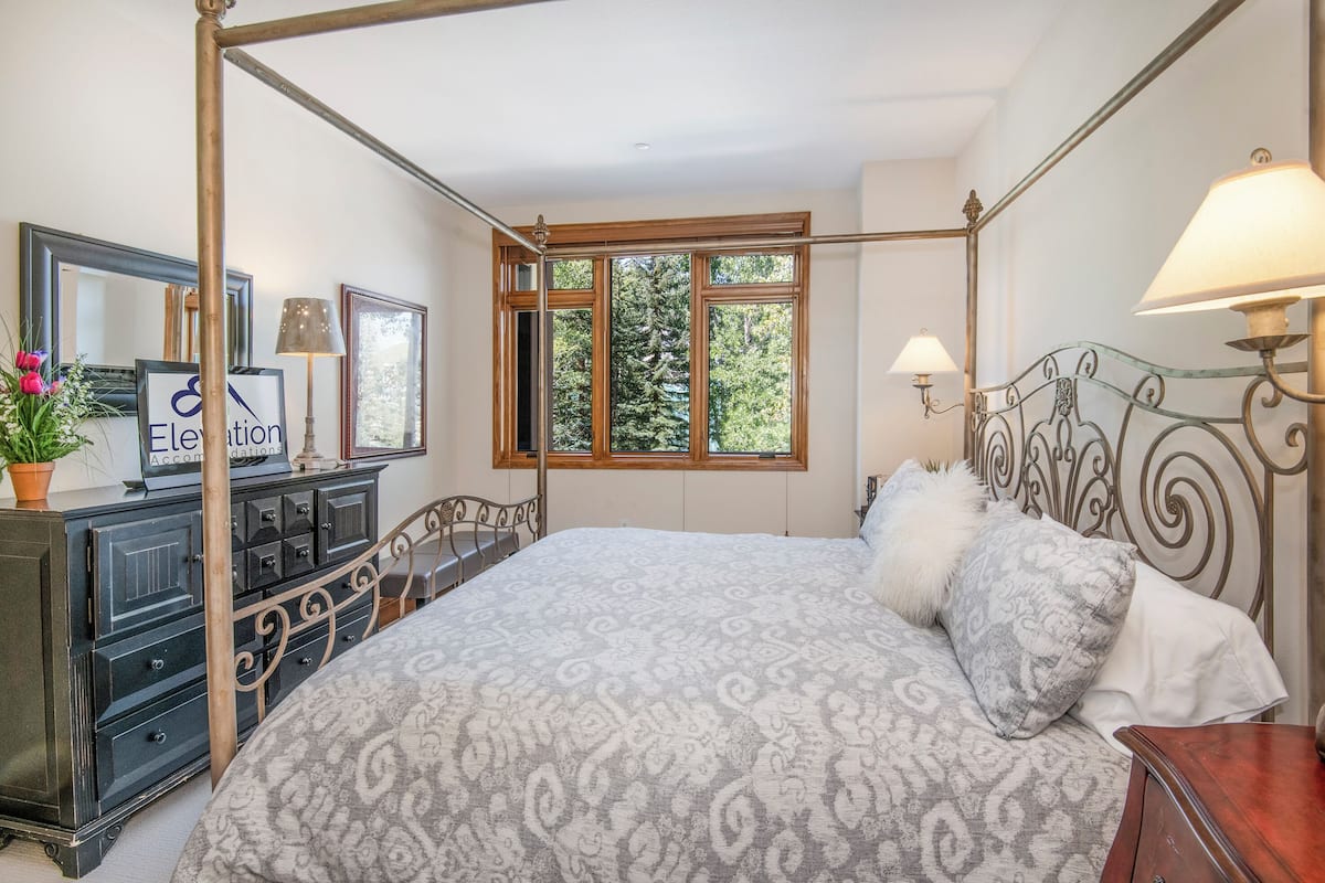 Un lit à baldaquin avec une couvre-lit à motifs, une commode avec un miroir et une fenêtre donnant sur des arbres.