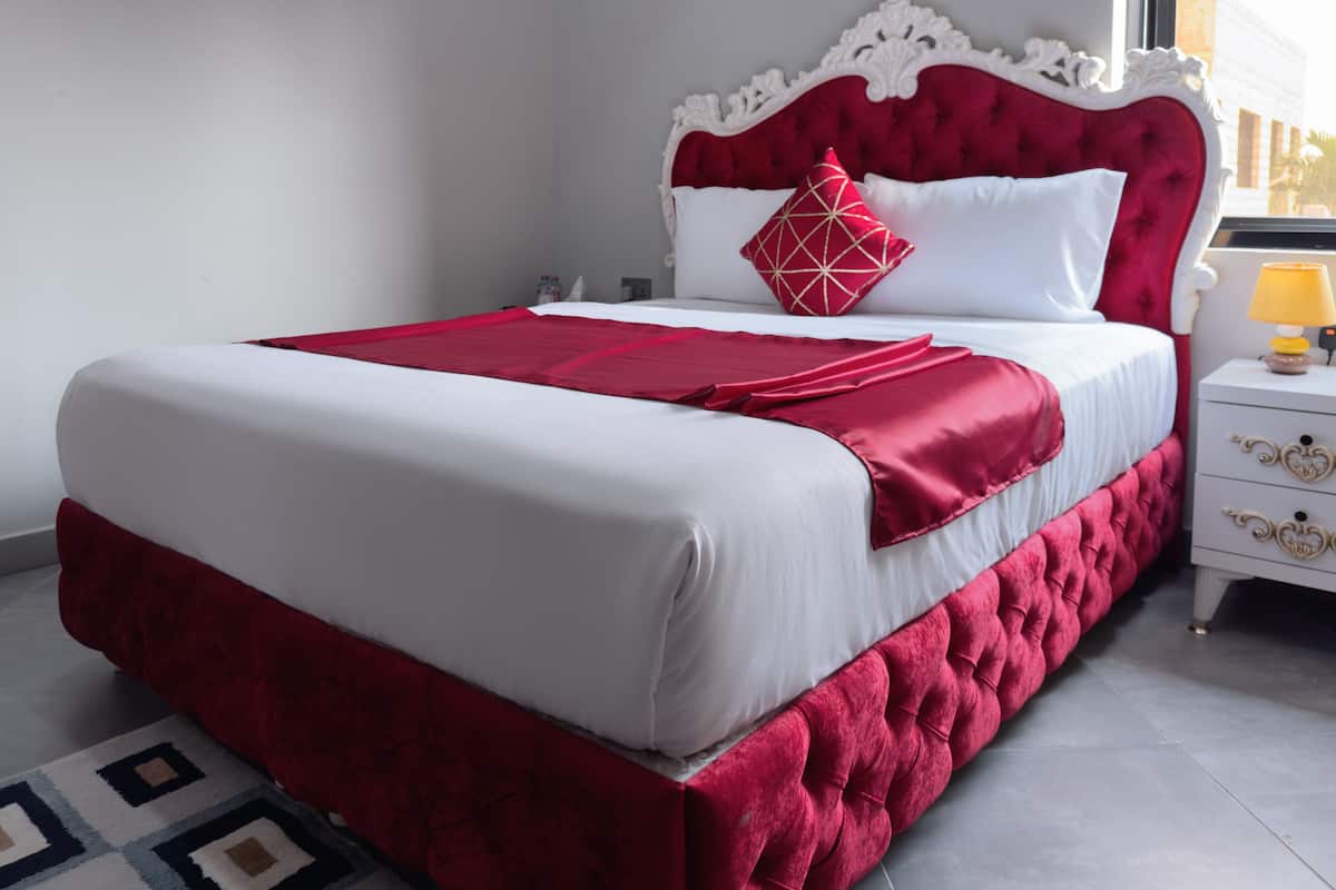 A neatly made bed with a red and white color scheme, a white headboard, and a bedside table with a lamp.
