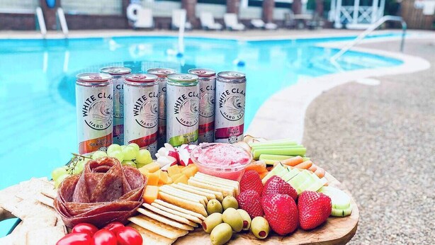 Ein Pool-Buffet mit verschiedenen Snacks und Getränken, darunter Dosen Hard Seltzer, Käse, Oliven, Erdbeeren und Aufschnitt.