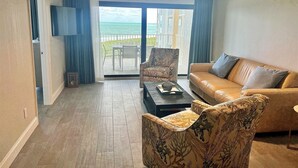 Ein Wohnzimmer mit Sofa, Sessel und Couchtisch, einem Deckenventilator und Blick auf das Meer durch eine Glastür.
