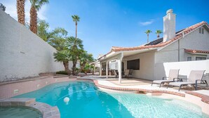 Une piscine avec un jacuzzi, entourée de palmiers et dotée d’un patio couvert avec des chaises longues.