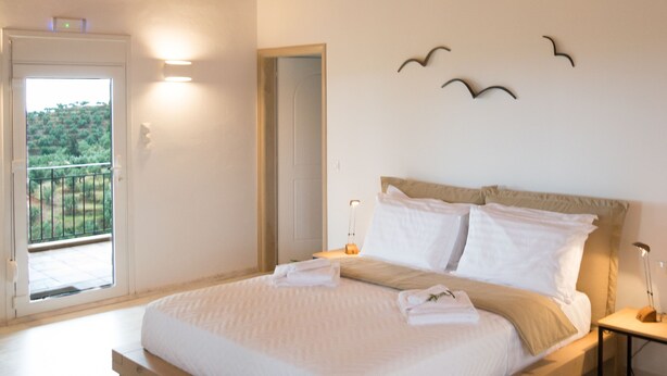 A bedroom with a wooden ceiling, a bed with white linens, a bedside table with a lamp, and a view through a door to the outside. - Olea Escape - Your own house in an exclusive olive estate | Stunning 360° Views (Githio)