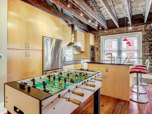 A modern kitchen with a foosball table, stainless steel appliances, and exposed brick walls.