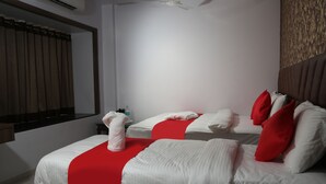 A hotel room with two beds, each with white bedding and red accents, a mirror, a ceiling fan, and a wall-mounted shelf with toiletries. - Hotel Apple Inn (Navi Mumbai)