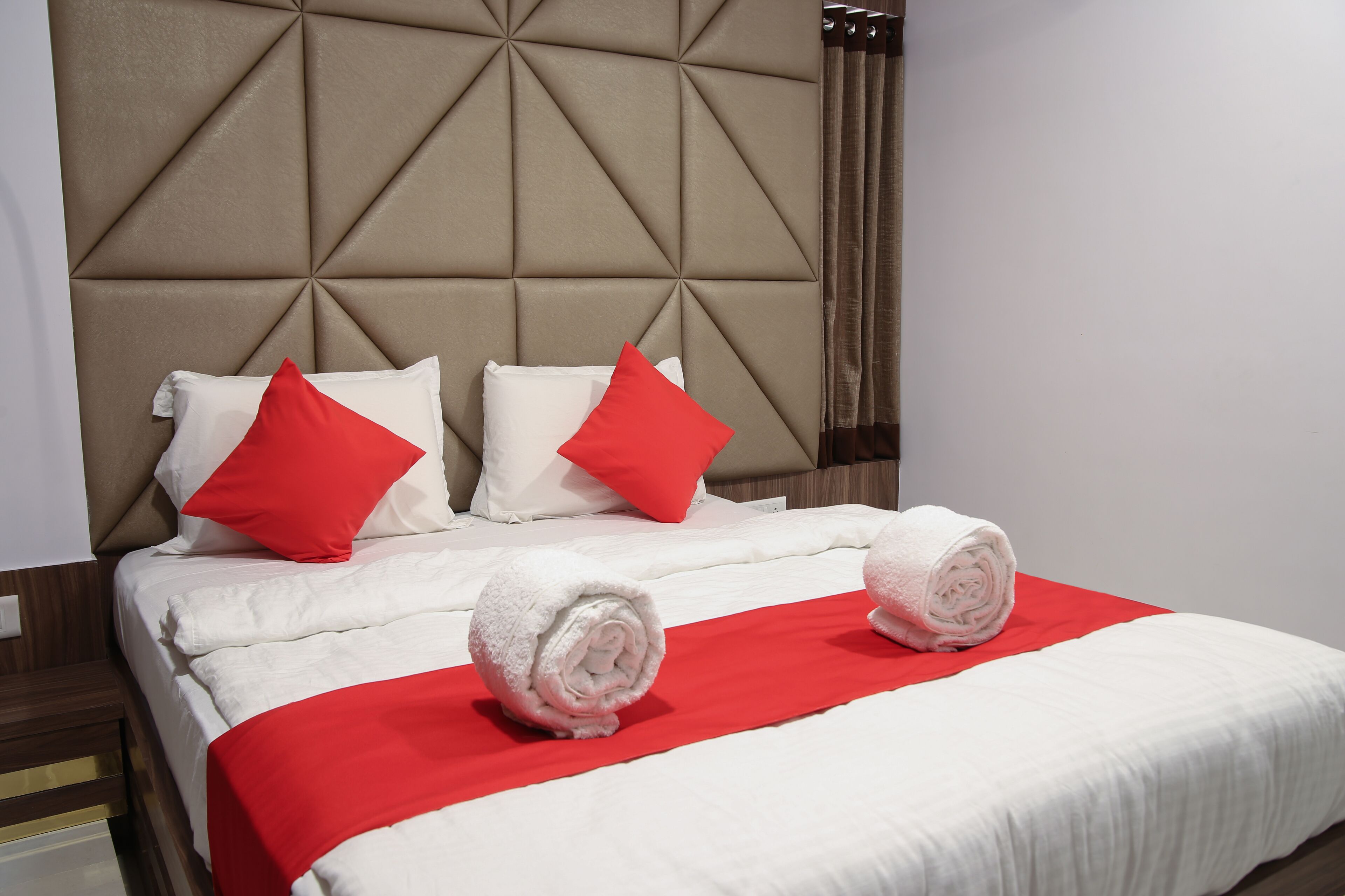 A hotel room with a tufted headboard, white bedding, red pillows, and rolled towels on a red runner.