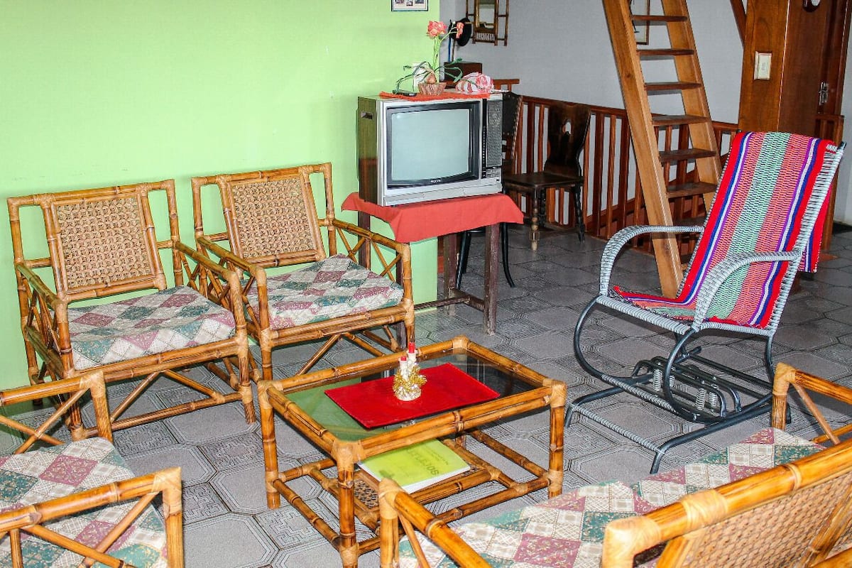 Een kamer met een televisie op een standaard, twee bamboe fauteuils met kussens, een kleurrijke geweven stoel, en een rode mat op de vloer.