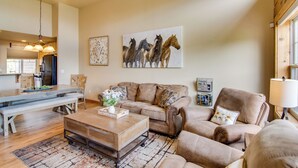 Un salon spacieux avec un grand canapé, une table basse et un coin repas avec un banc. Un tableau représentant des chevaux orne le mur, et on aperçoit une cuisine au fond.