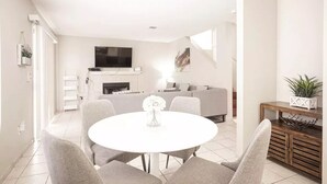 Een moderne woonkamer met een witte plafondventilator, een flatscreen-televisie boven een open haard, een grijze bank en een eethoek met een ronde tafel en grijze stoelen.
