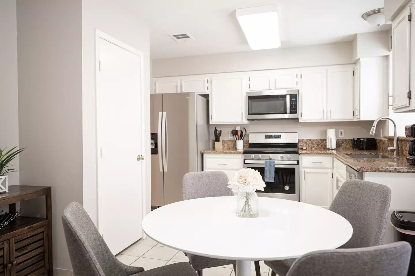A modern kitchen with white cabinets, a microwave, oven, and a sink. There is a dining table with four chairs and a vase with flowers.