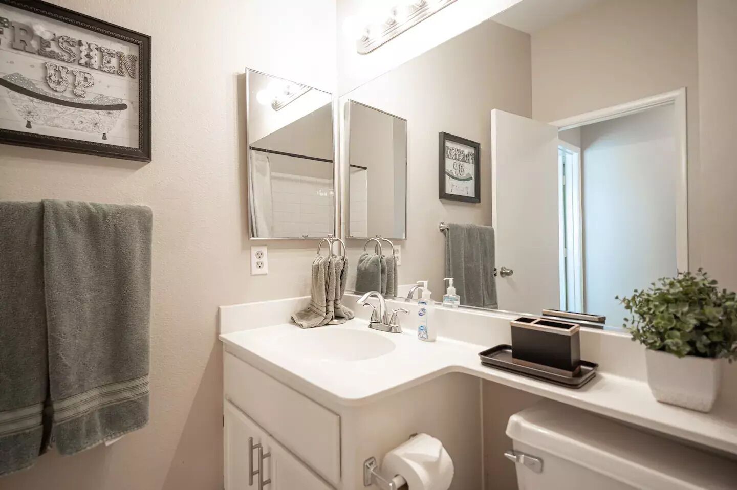 A bathroom with a white sink, two mirrors, a towel rack, a toilet, and a framed picture on the wall.