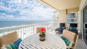 Een balkon met een ronde tafel, twee stoelen, een plant in pot en uitzicht op het strand en de oceaan.