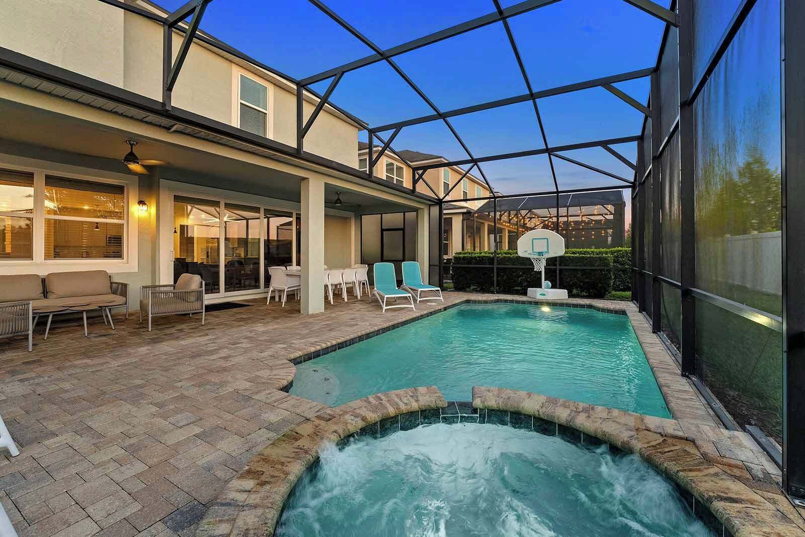 An outdoor swimming pool with a hot tub, surrounded by a covered patio area with seating and a basketball hoop.