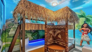 Una stanza con un letto a tetto di paglia, un murale raffigurante una scena tropicale e un’illustrazione di un personaggio.