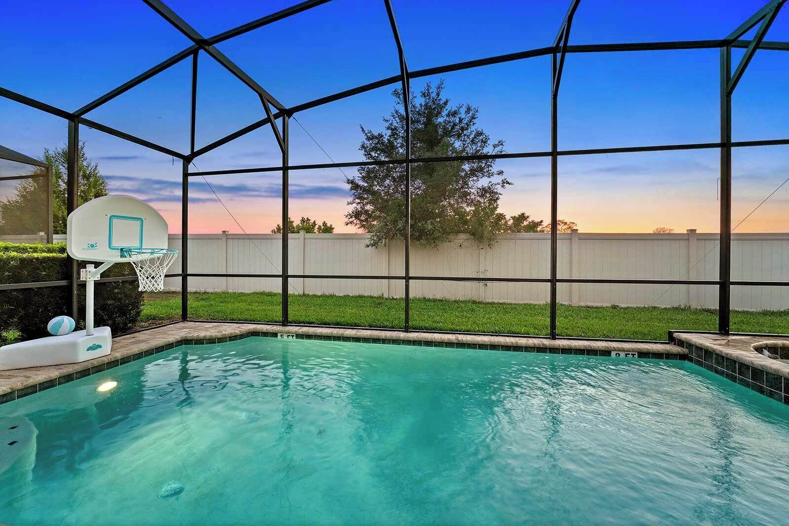 A swimming pool with a basketball hoop and a view of a white fence and trees.