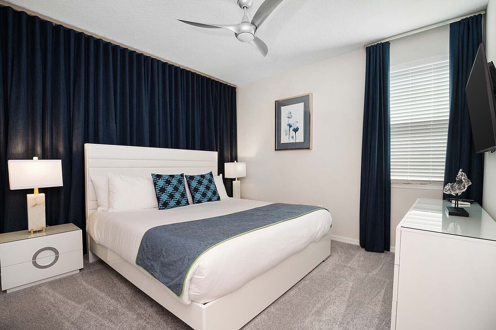 Une chambre à coucher bien rangée, avec un lit, des lampes de chevet, un ventilateur de plafond, un tableau encadré au mur et une fenêtre avec des rideaux.