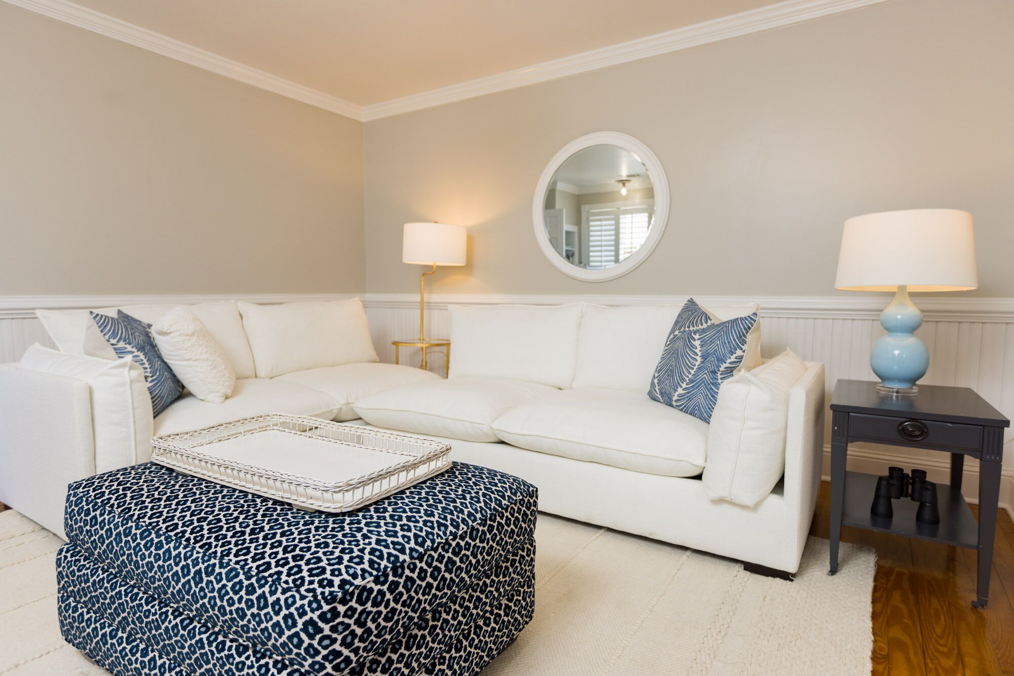 Un salon avec un canapé blanc, un miroir rond, deux lampes et un pouf à motifs.