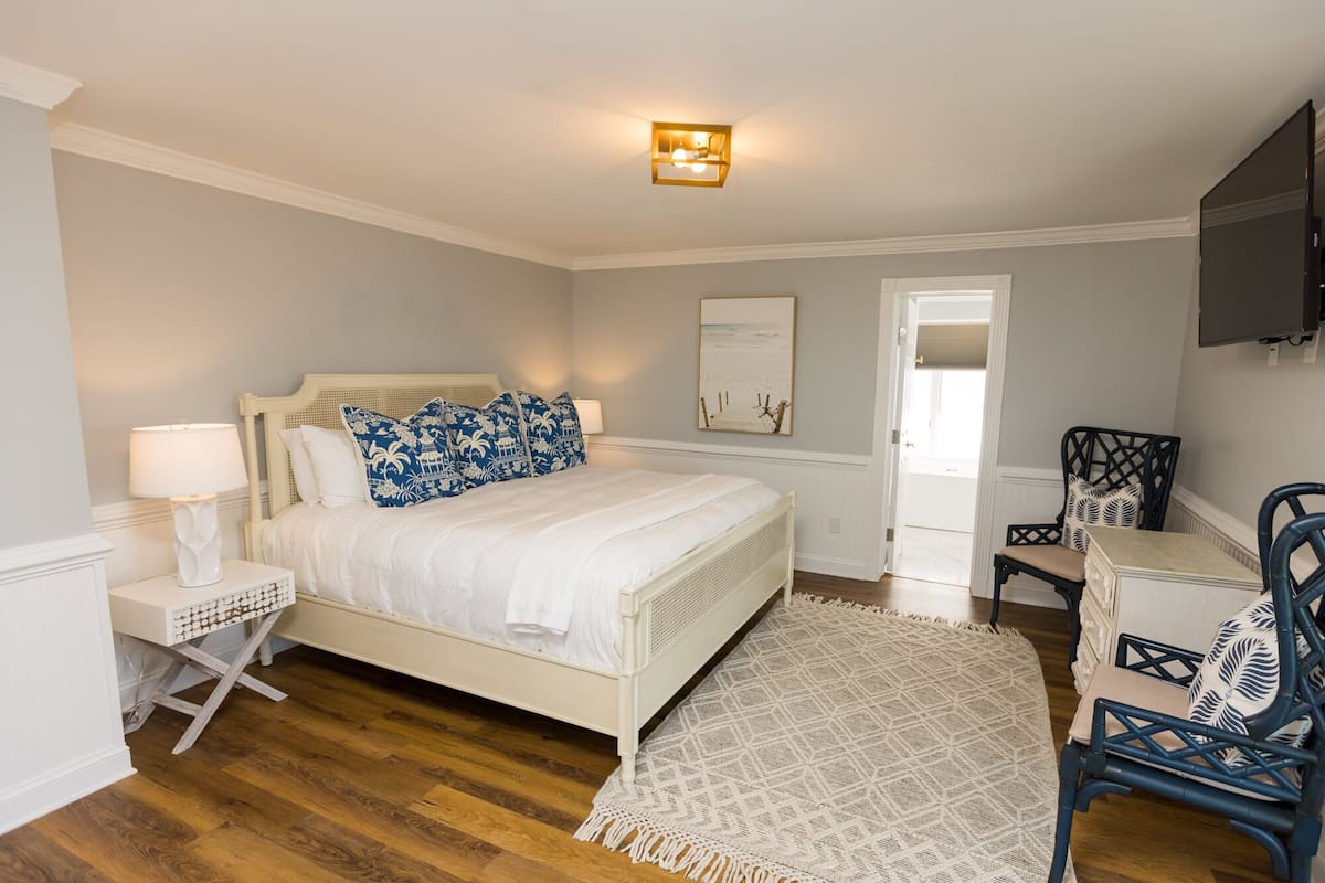 A bedroom with a white bed, a bedside table with a lamp, a chair, a desk, and a painting on the wall.