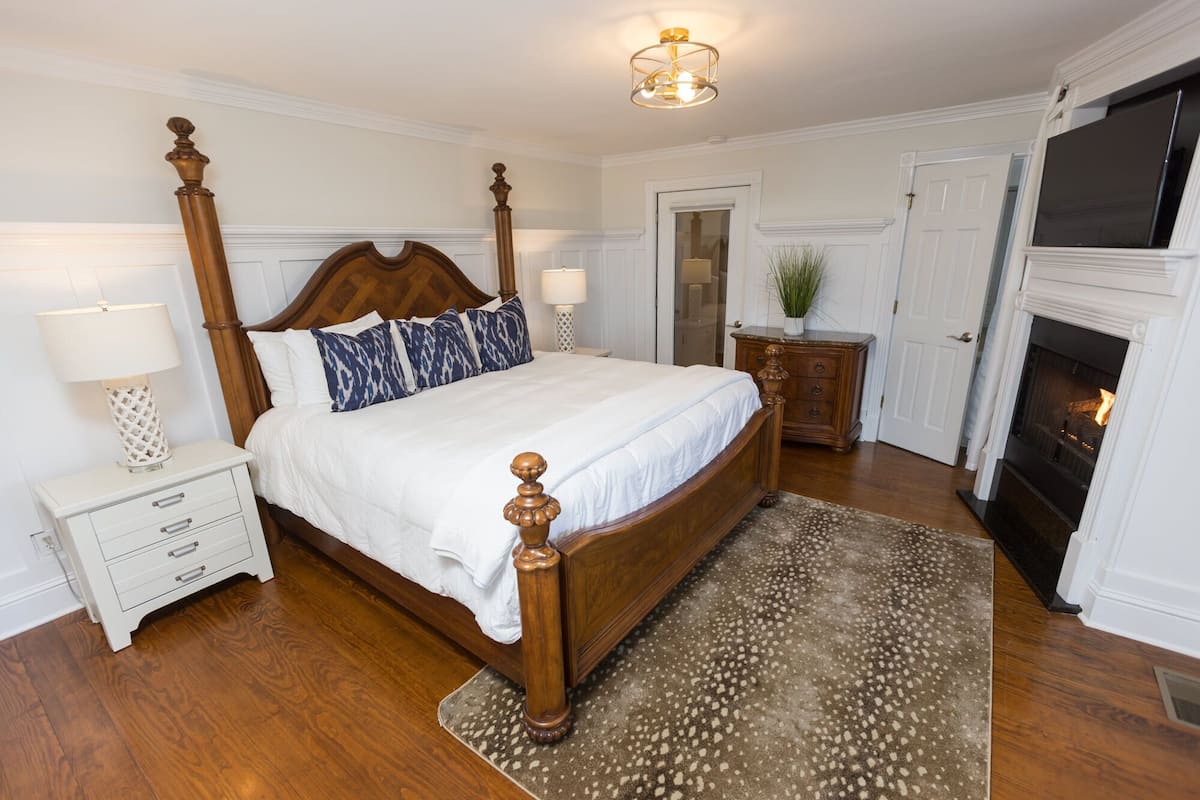 A bedroom with a four-poster bed, a nightstand with a lamp, a dresser, a mirror, and a fireplace.