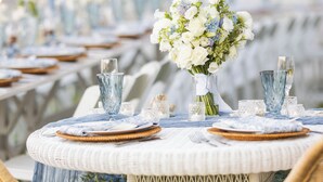 A round table with a white wicker tablecloth, blue and white floral arrangements, and glassware. - The Burrus House Inn Waterfront Suites (Manteo)