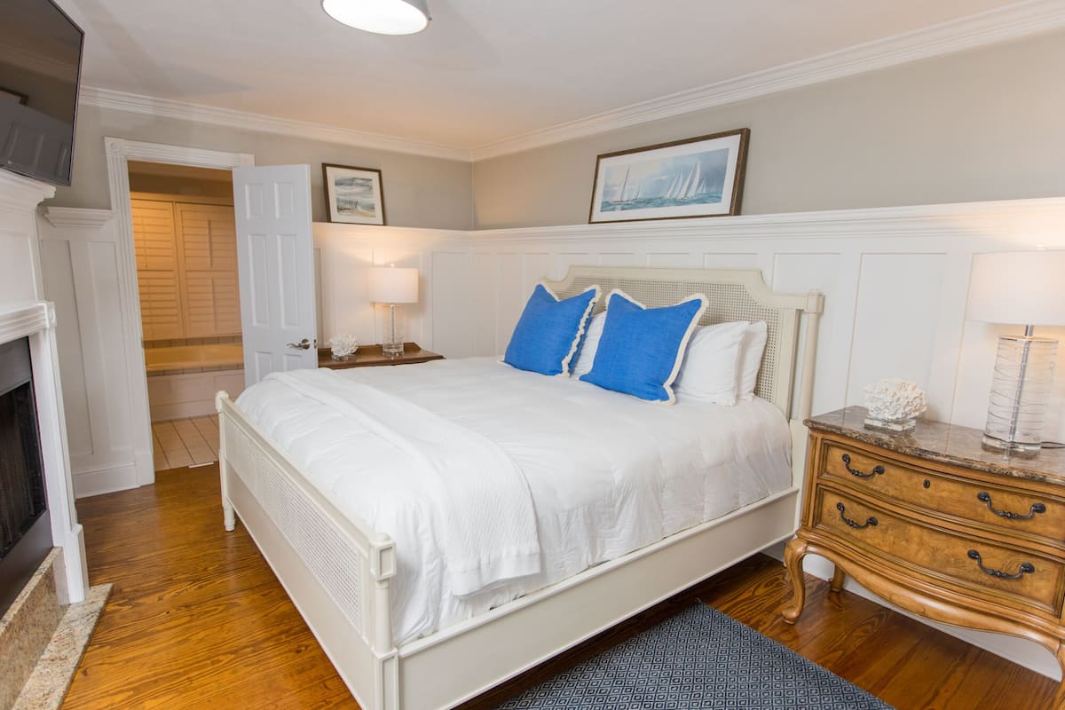 A bedroom with a white bed, blue pillows, a wooden nightstand, and a fireplace.