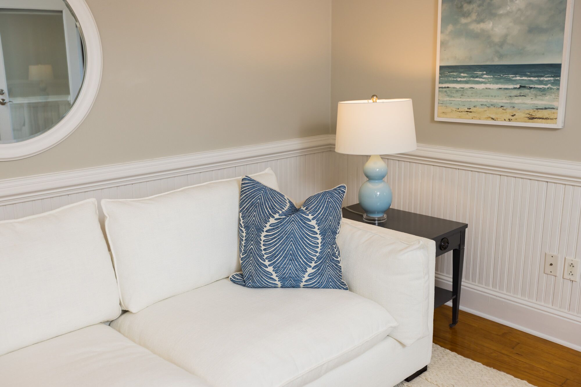 Ein weißes Sofa mit einem blauen Kissen mit Muster, ein kleiner Tisch mit einer Lampe und ein Gemälde einer Strandansicht an der Wand.
