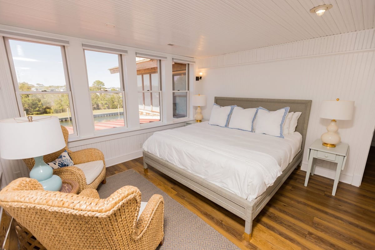 A bedroom with a large bed, two bedside tables, a wicker chair, and a view of a pool through the windows.