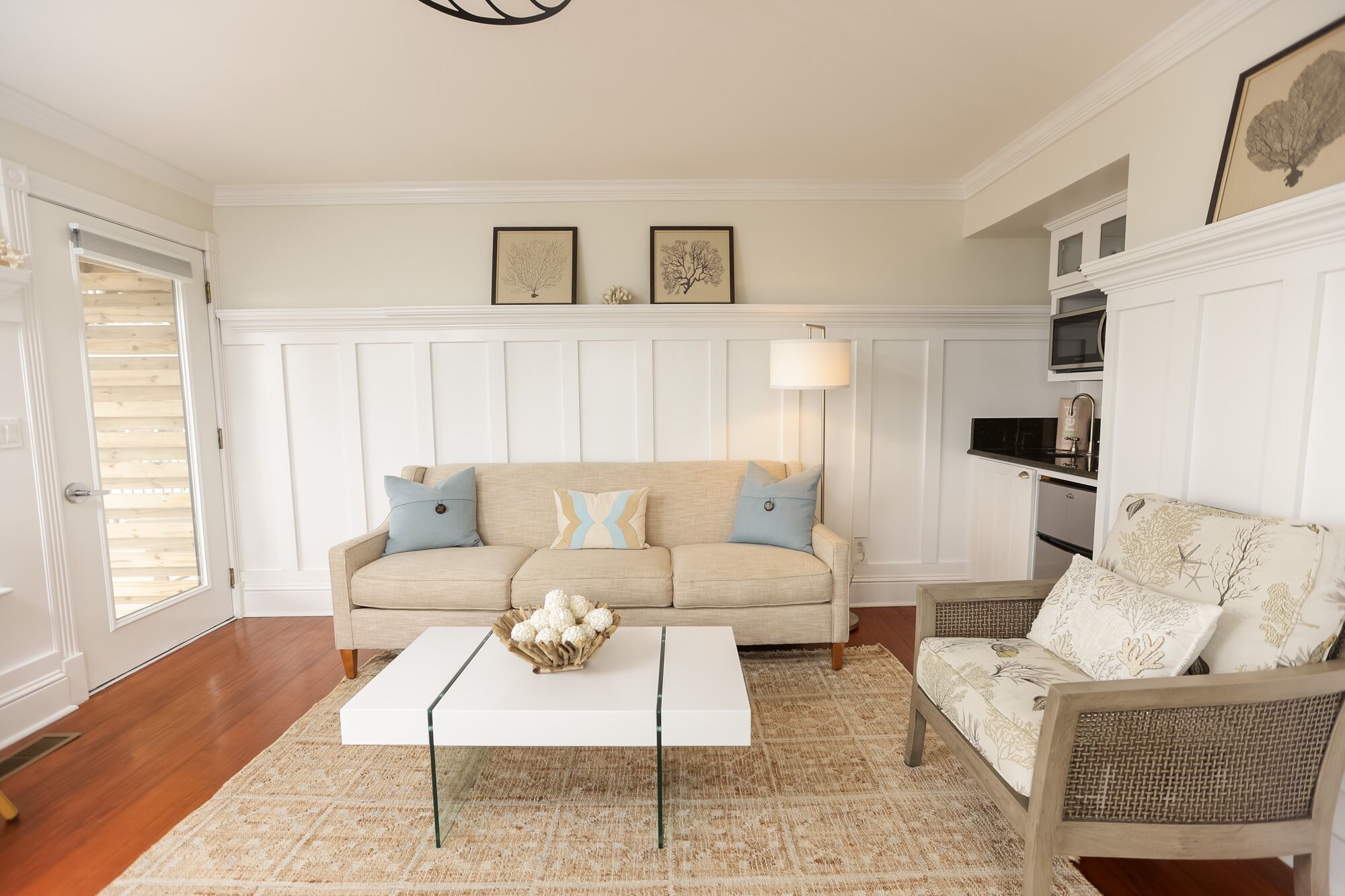 Een woonkamer met een beige bank, een witte salontafel en een rieten fauteuil. Boven de bank hangen twee ingelijste foto's.