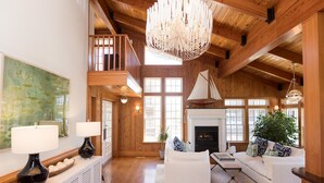 A spacious living room with a high ceiling, wooden beams, a fireplace, and a large window. - The Burrus House Inn Waterfront Suites (Manteo)