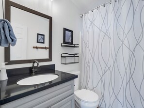 Une salle de bain avec un lavabo au comptoir noir, des toilettes blanches, un rideau de douche à motifs géométriques et un miroir avec un cadre en bois.
