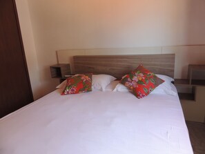 A neatly made bed with white linens and two floral pillows, a wooden headboard, and a nightstand with a remote control. - Our House in Jeri 2 (Jijoca de Jericoacoara)