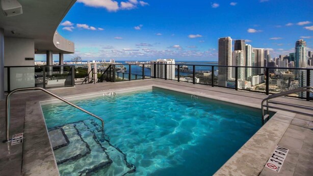 Ein Pool auf dem Dach mit klarem, blauem Wasser, umgeben von einem Metallgeländer, mit Blick auf eine Stadtlandschaft mit hohen Gebäuden und den Blick auf das Meer.