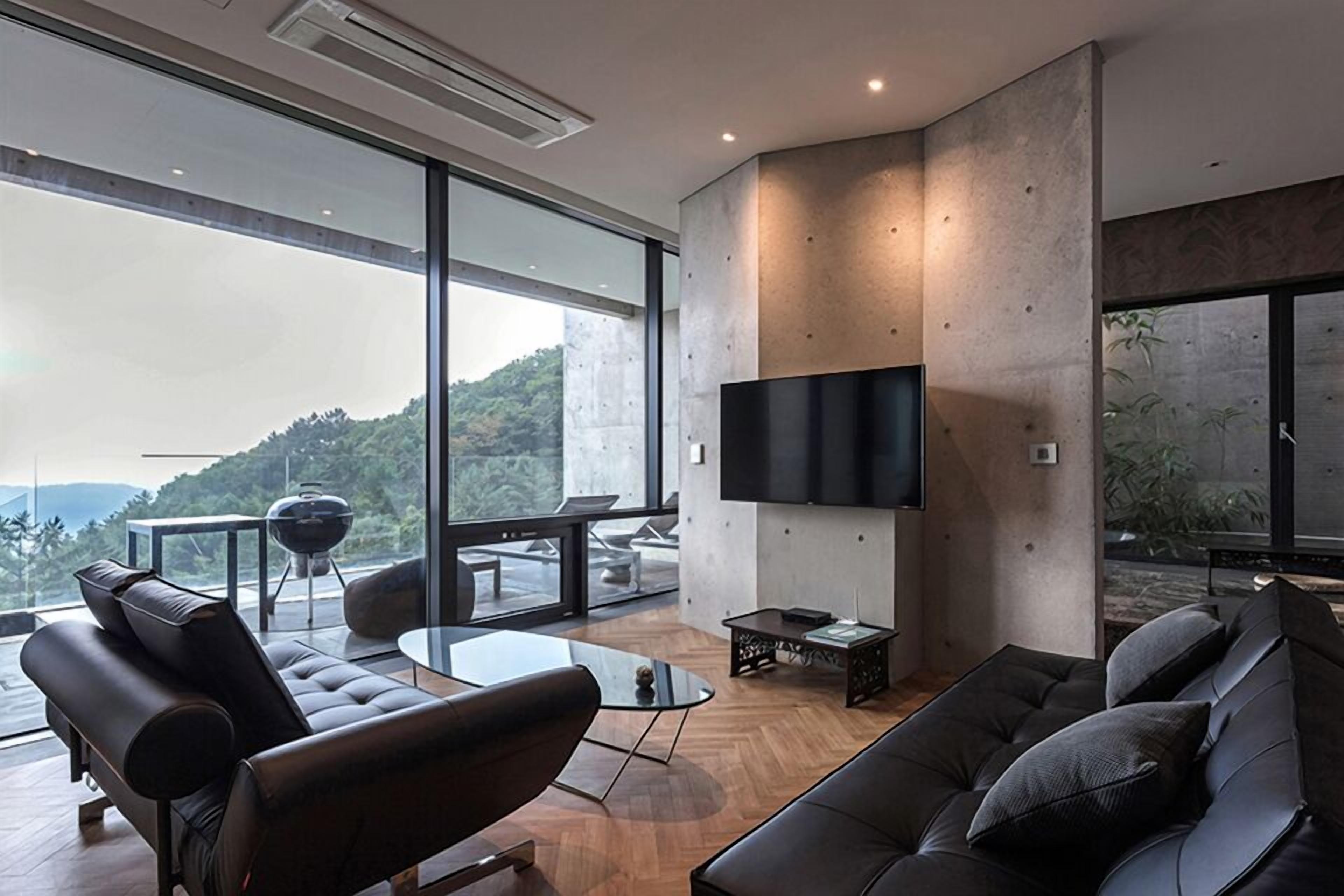 A modern living room with a large TV, comfortable seating, and large windows offering a view of greenery.
