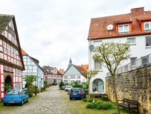 Une rue pavée bordée de maisons traditionnelles à colombages, de voitures garées et d’un mur de pierre sur lequel est adossé un banc.