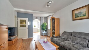A living room with a grey sofa, wooden floor, a wooden cabinet, a television, a bench with a purple cloth, and a painting on the wall.